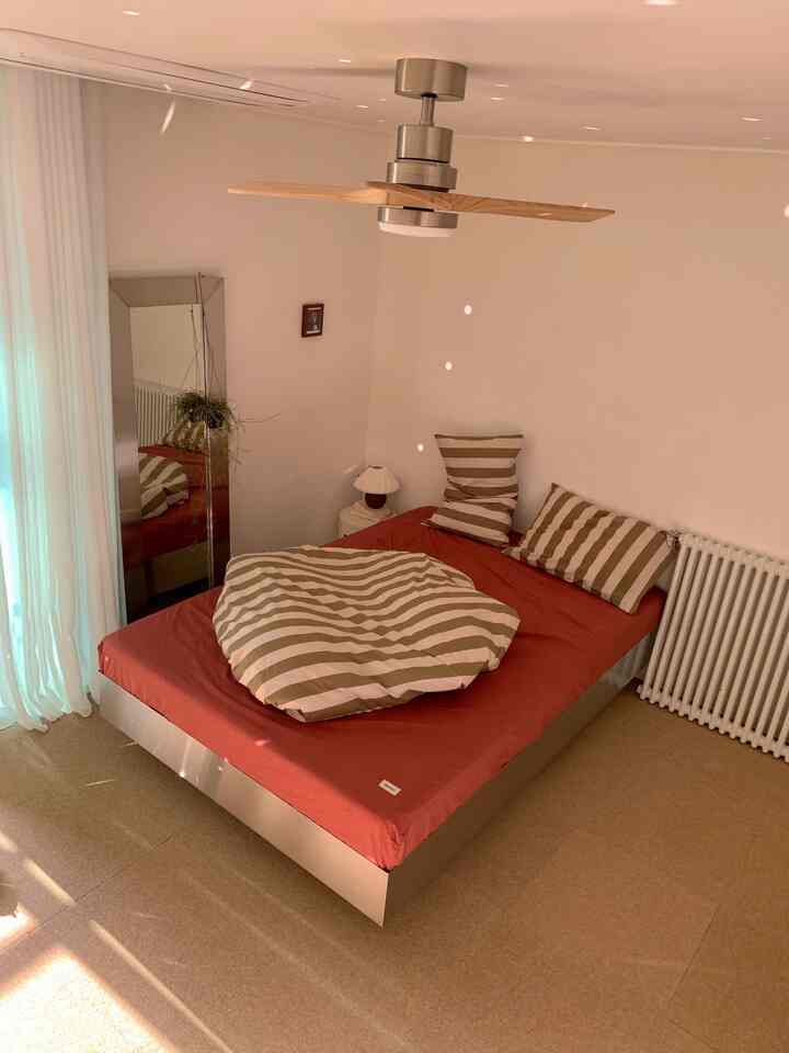 白い壁と茶色の床が調和する寝室、中央にベッドとストライプの掛け布団が配置された落ち着いた空間