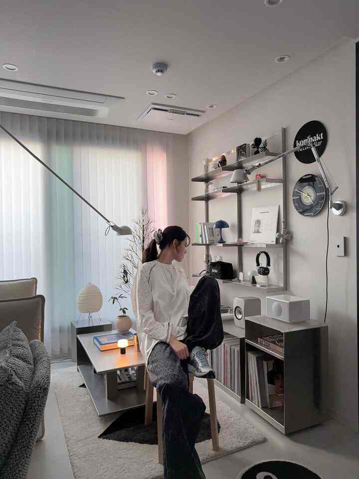 Bright white and gray toned living room featuring a simple modern sofa, aluminum coffee table, wall shelves, and wall lighting