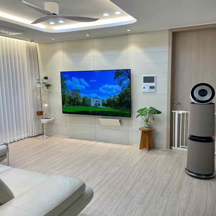 ホワイトトーンのミニマルなリビング、透明な本棚や壁掛けテレビ、空気清浄機、ソファが配された清潔感ある空間