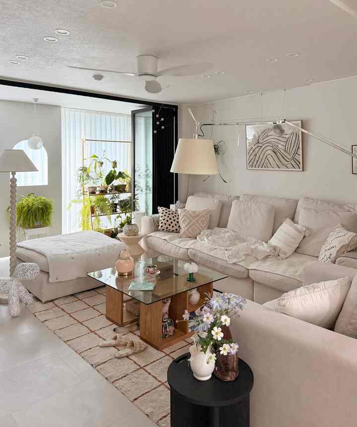 明るいホワイトとベージュ調のリビング、クリーム色のコーナーソファとガラスのコーヒーテーブル、植物棚、多様なクッション、フロアランプが配置された自然で温かみのあるインテリア