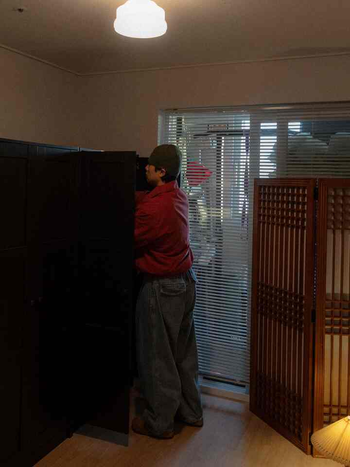 暗い家具とアルミブラインドがある小さく落ち着いた部屋でワードローブを開ける人