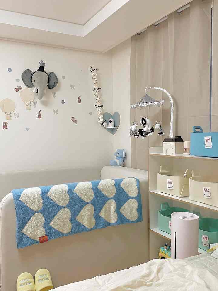 ホワイトとベージュの色調の子供部屋、ベビーベッドとモビール、ブルーのハート柄スローがあるシンプルな空間