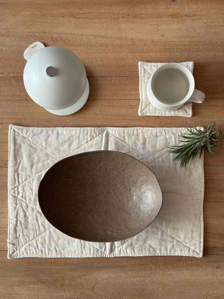 ナチュラルな木目調のキッチンテーブルに、茶色い皿と白いマグカップ、コットンマット、空気清浄の植物が配置された清潔で自然な雰囲気のインテリア