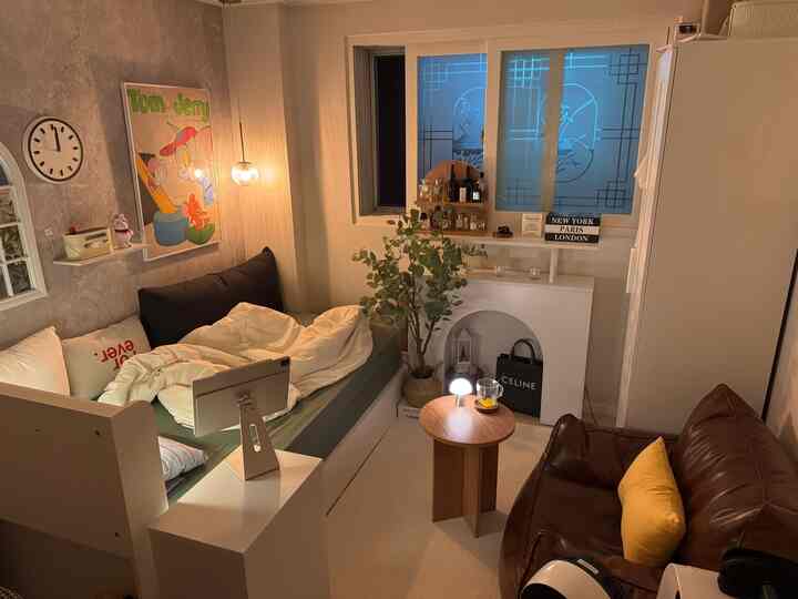 White and brown toned small studio featuring a cozy bed, sofa, fireplace console, and warm natural modern interior lighting