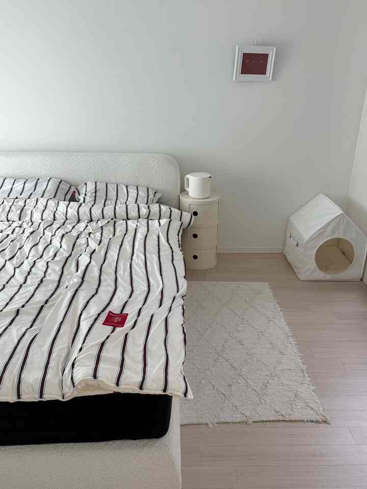 白とナチュラルカラーの寝室、中央左にストライプ柄のベッド、右側にペットハウスが配置されたミニマルで落ち着いたインテリア