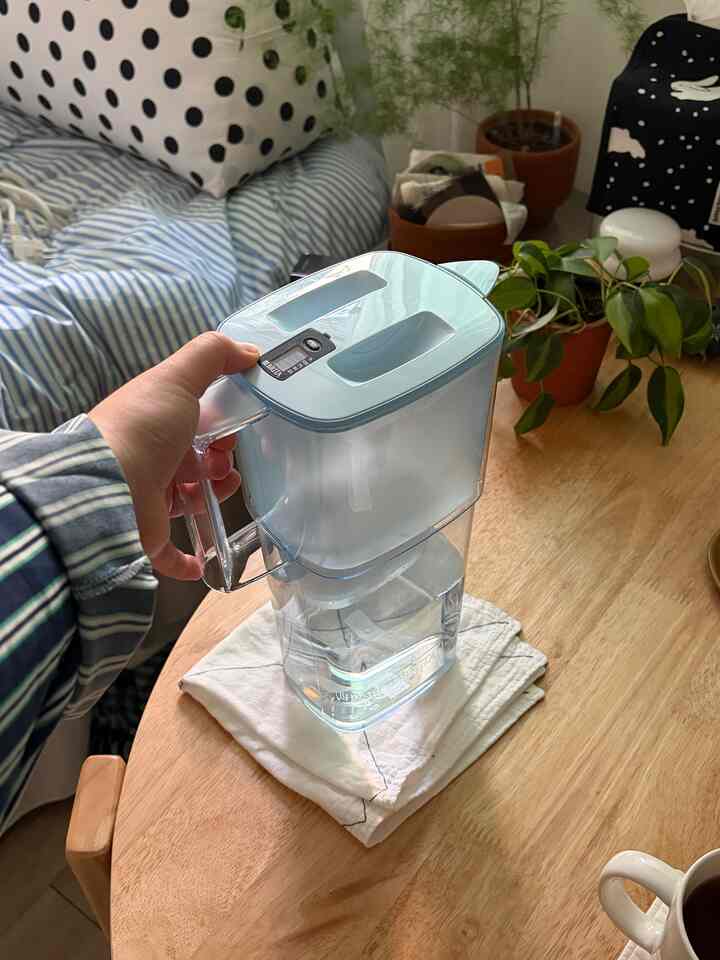 ナチュラルな色調のキッチンダイニングテーブル、ブリタの浄水器を持つ手、鉢植えとクッションが配置された雰囲気のある空間