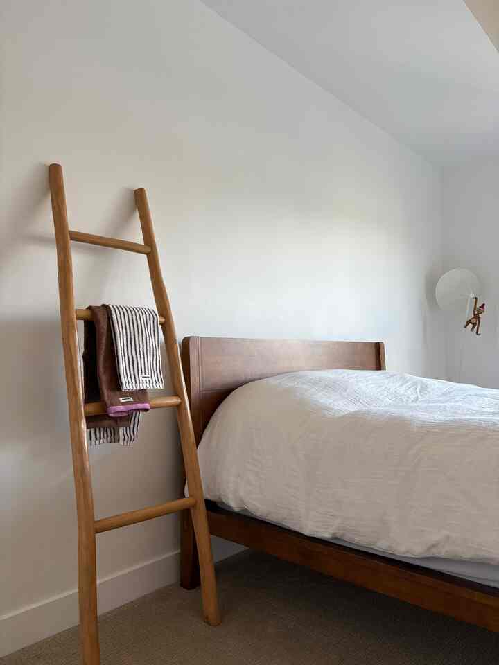Natural wood tone bedroom with white walls and bedding, featuring wooden ladder holding towels for a cozy atmosphere