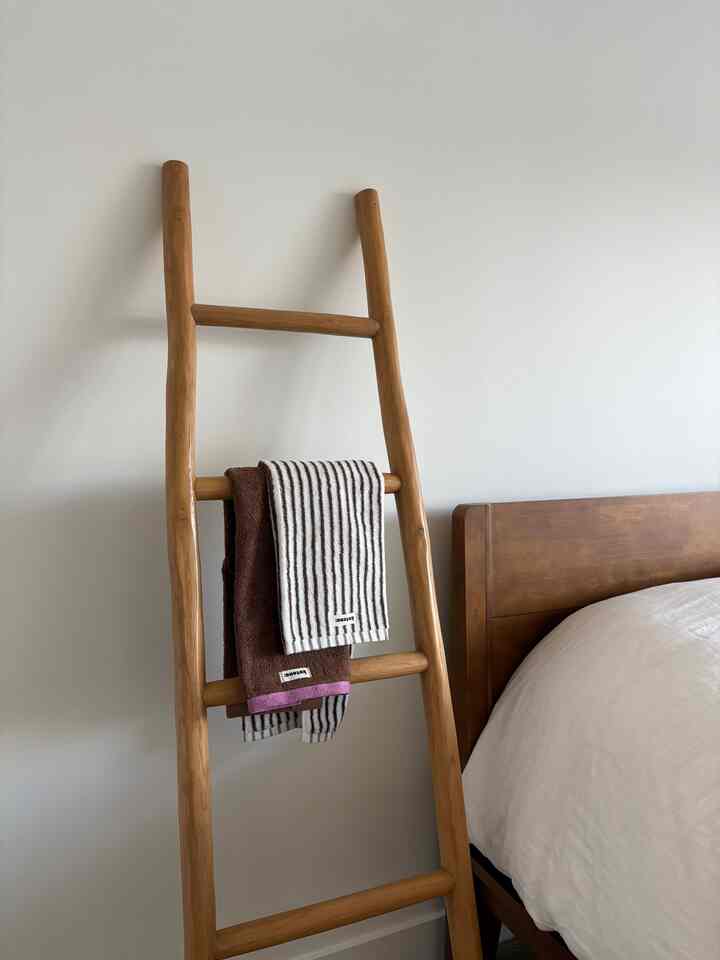 Wood tone leaning wooden ladder with towels hanging beside a wooden bed in a simple bedroom space