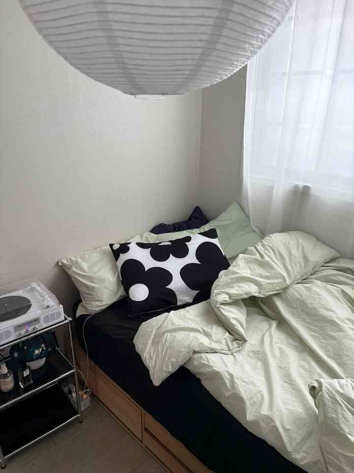 明るいベージュ調の寝室、ブラックのマットレスと花柄のクッション、収納付きの木目調ベッドが配置された居心地の良い空間