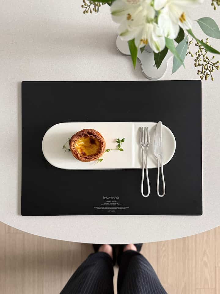 ホワイトのテーブル上にブラックのロウバックテーブルマットが置かれ、ナチュラルな色合いの長楕円形プレートにデザートとカトラリーが美しく配置された雰囲気のあるインテリア