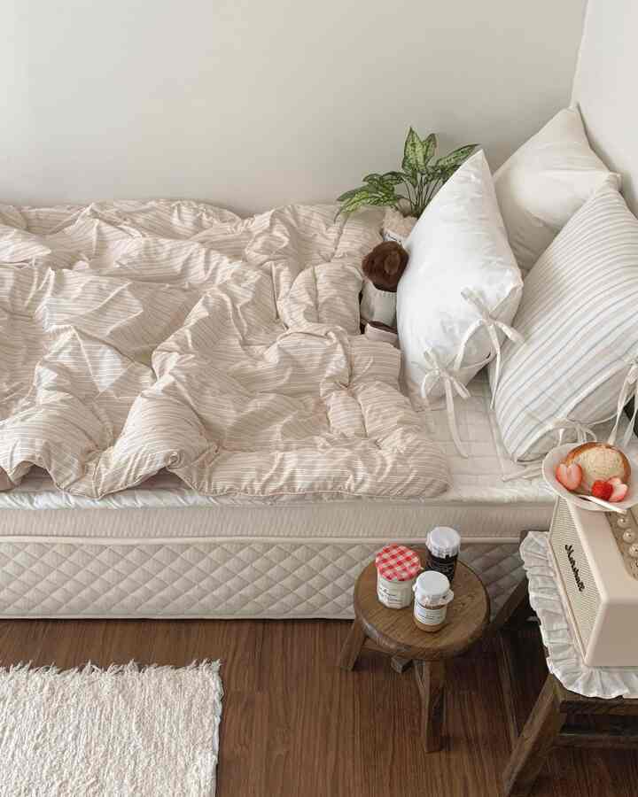 A beige and white toned bedroom featuring a queen-size bed with mattress, pillows, and wooden stools creating a cozy atmosphere