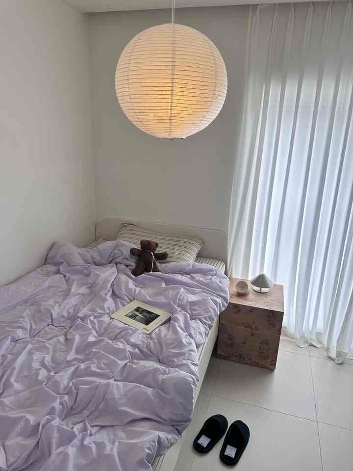 White-toned kids' bedroom featuring soft lavender bedding, a natural wood stool, and a round paper pendant lamp creating a cozy atmosphere