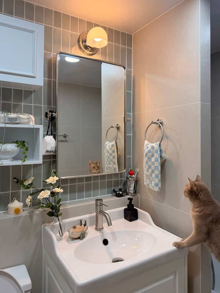 ホワイトとグレーのタイルが配されたバスルーム、白い洗面台と鏡、タオルや造花が配置されたヴィンテージ感のあるインテリアに猫がいる