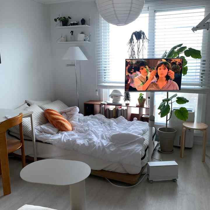 White and natural toned studio bedroom featuring a bed, bookshelf, and floor lamp with a cozy atmosphere