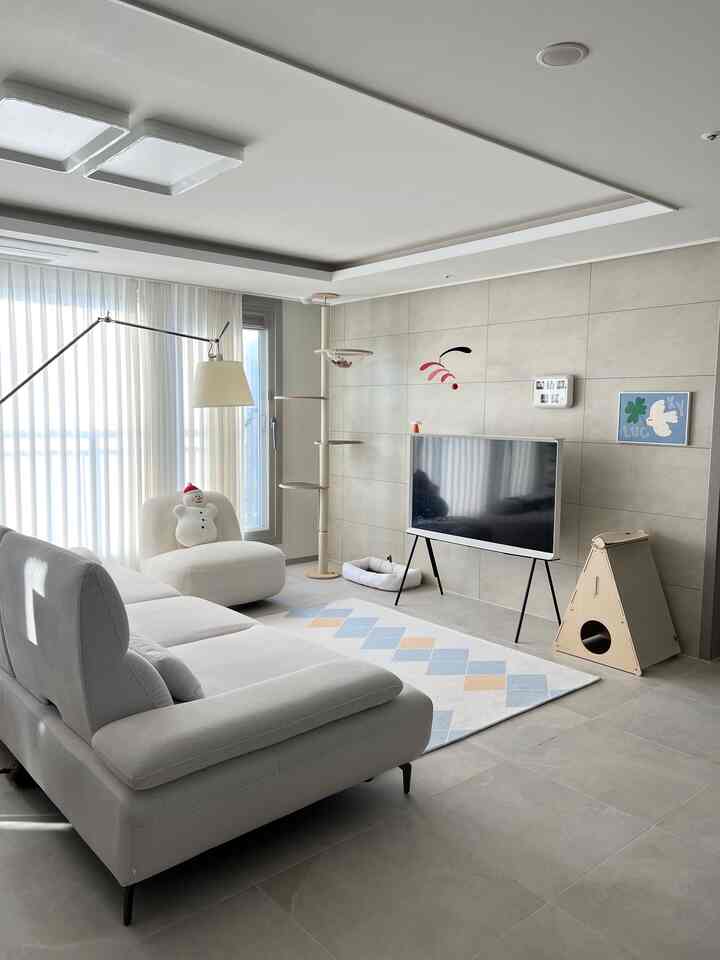 明るいナチュラルトーンのリビング、大きなソファとキャットタワー、ラグ、テレビが配置された落ち着いたモダンな空間