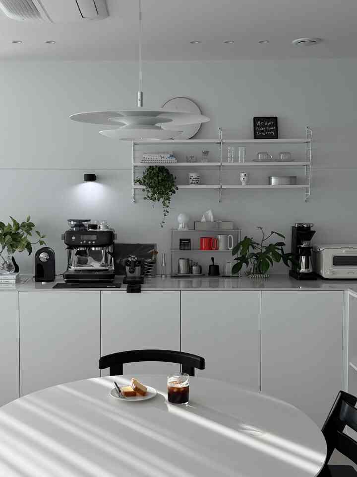 ホワイトトーンのキッチンスペース、モダンなテーブルとコーヒーマシン、棚、植物が配置されたミニマルな印象的なホームカフェインテリア