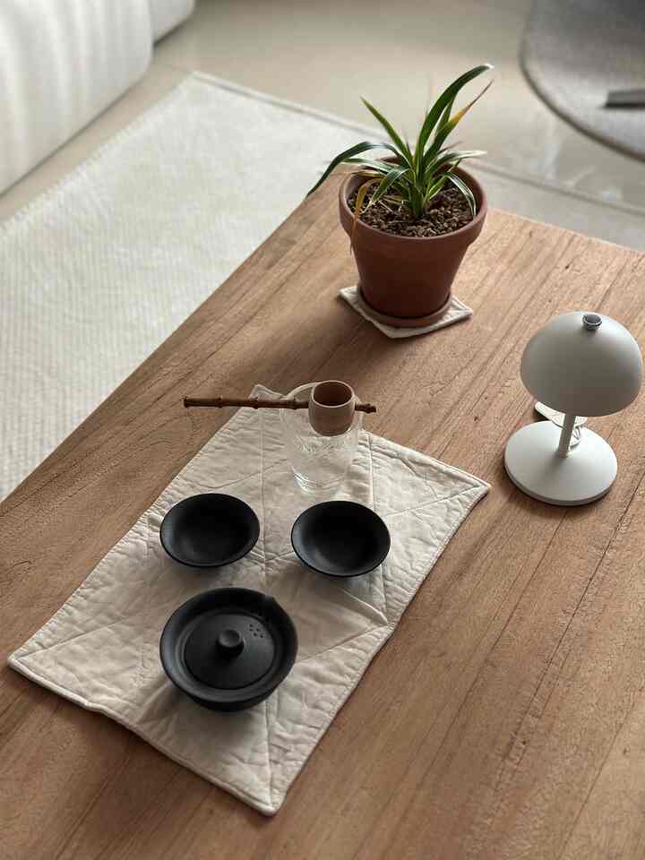 柔らかなブラウンの木製テーブルの上に黒い茶器セットと白いランチョンマット、小さな植物と白いテーブルランプが配置された温かみのあるインテリア