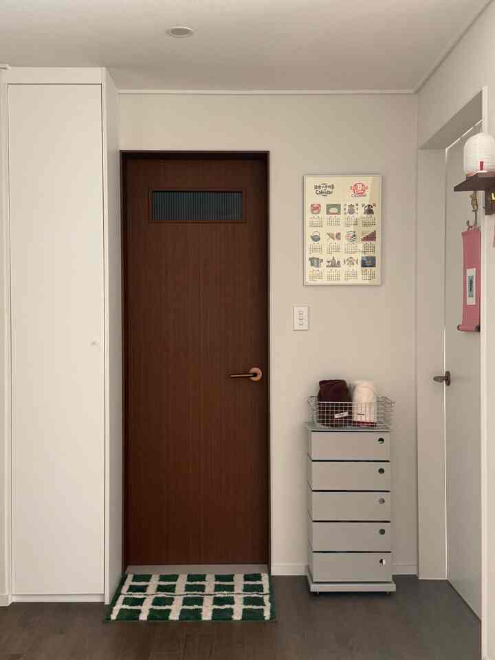 白い壁と木目調の扉が調和したシンプルなバスルーム入口、収納チェストとタオルバスケットが配置されたミニマルな空間