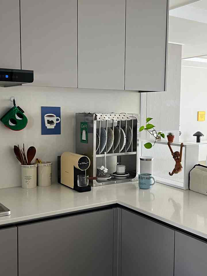 白とグレーのトーンのキッチンコーナー、カプセルコーヒーマシンとステンレスのラック、小さな植物が配置された落ち着いたおうちカフェの空間