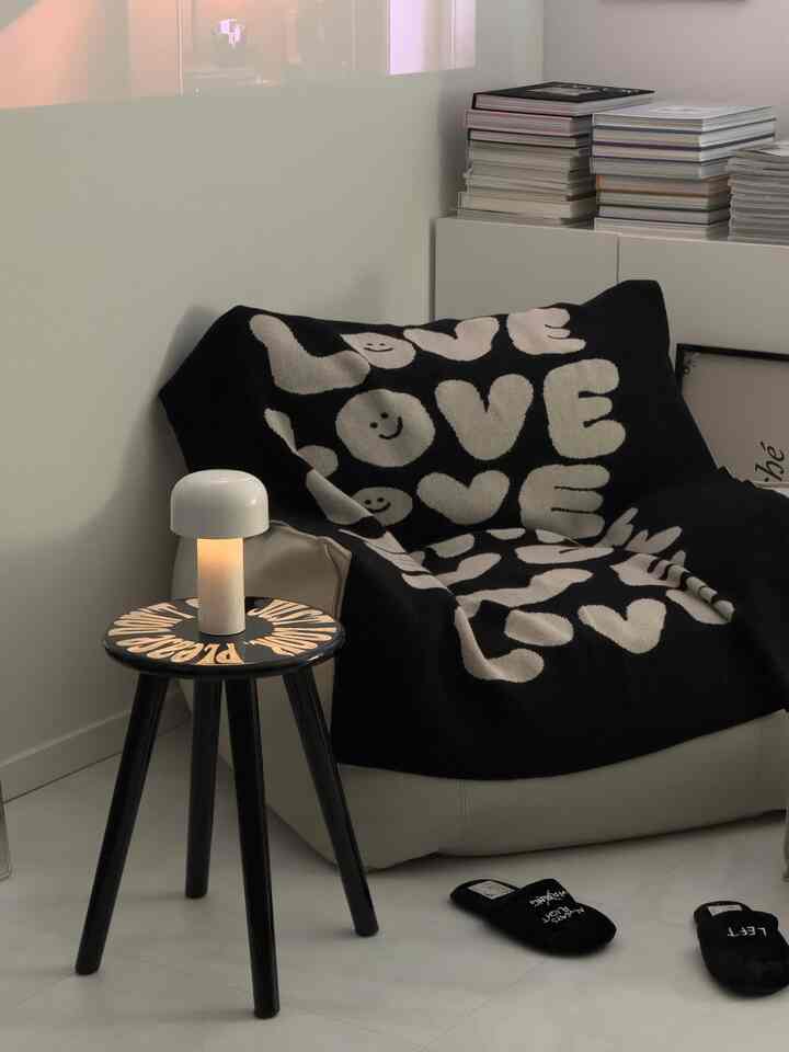 白と黒のトーンの一人暮らし空間、LOVE柄のブランケットと小さな黒いスツール、テーブルランプが置かれた落ち着いたモダンなインテリア