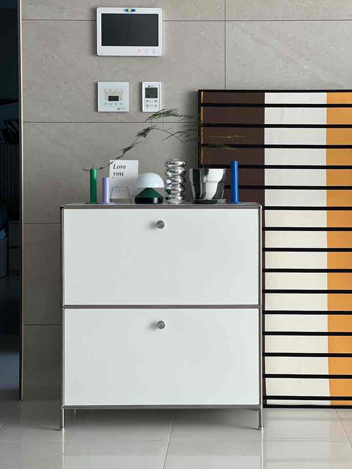 白いキャビネットの上にシルバーの花瓶と様々なオブジェが置かれたシンプルでナチュラルカラーのリビングスペース