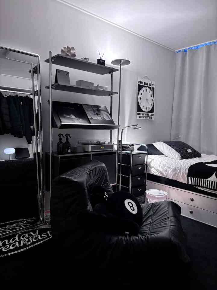 白い壁と遮光カーテンがあるミッドセンチュリーモダンスタイルのワンルーム寝室、黒い家具とLPプレーヤーが配置されたミニマルなインテリア