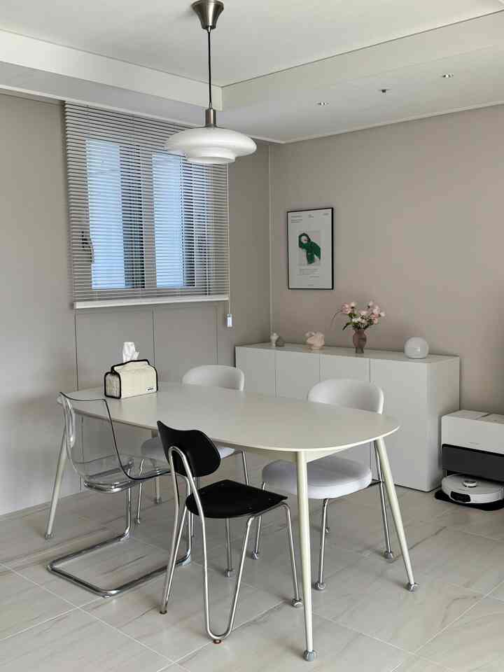 Minimal dining room with light beige walls and white furniture including dining table, accented by vase with flowers and transparent chair for a clean look