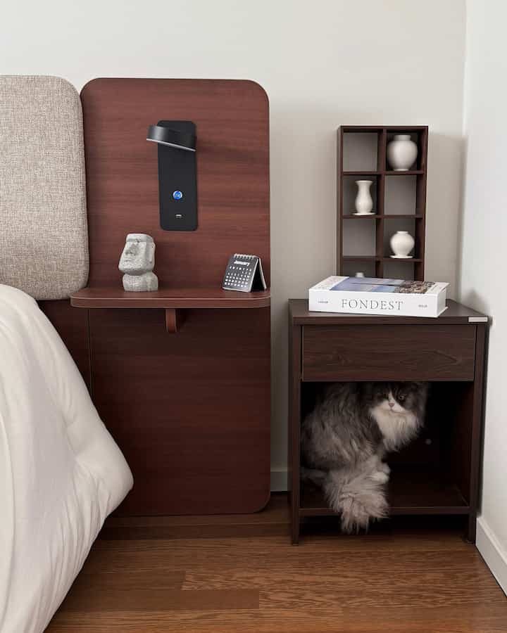 ブラウンのウォールナットのナイトテーブルとベージュの寝具が配置されたナチュラルな寝室、ナイトテーブルの下に座る猫が見える印象的な空間