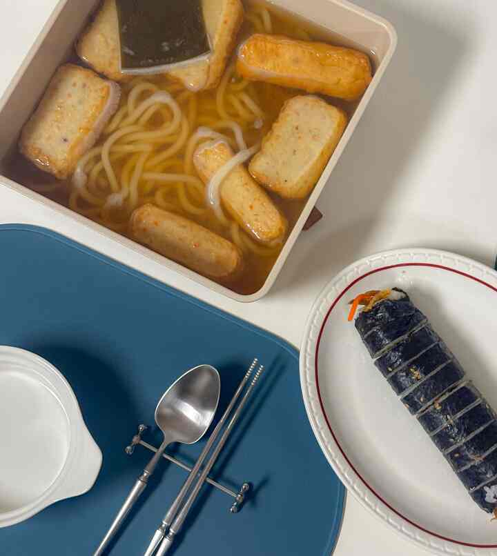 ベージュの鍋にうどんと練り物、白いテーブルの上にカトラリーとキンパが置かれたシンプルな食事空間