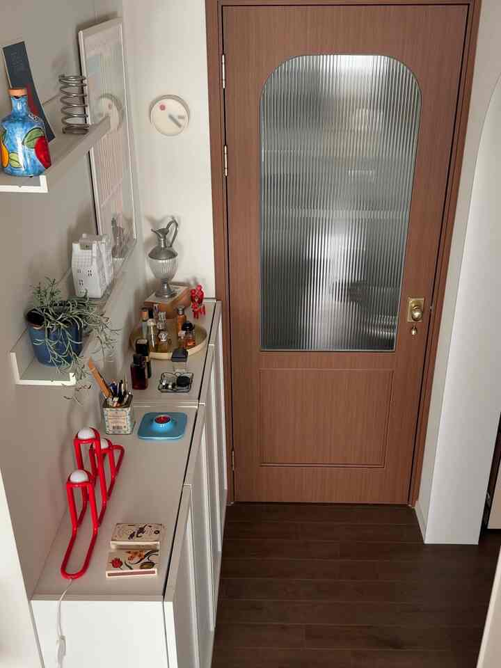 白い壁と家具、茶色のドアと床が調和する狭い玄関、棚と植物が配された雰囲気のあるインテリア