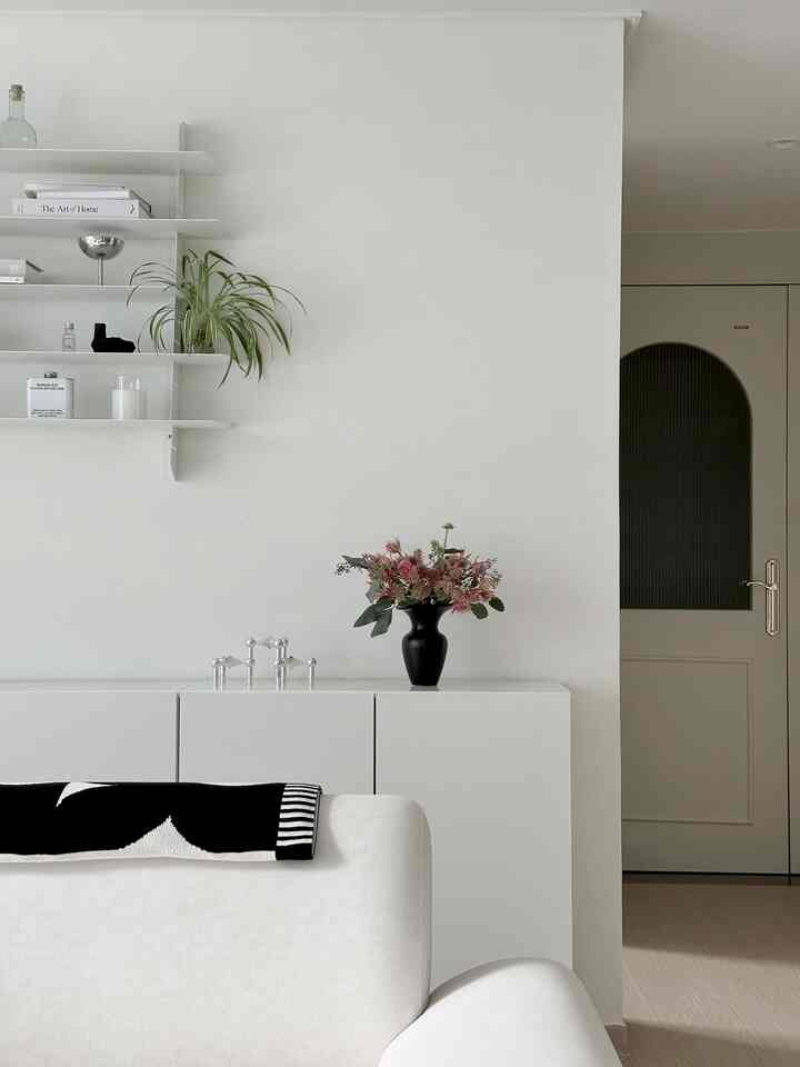 Monotone white living room featuring a cozy sofa, white shelves, a black vase with flowers, and green plants in a simple interior space