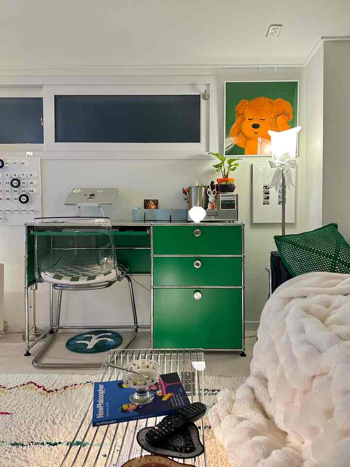 Natural-toned home office featuring a green metal desk, transparent chair, patterned rug, plants, and a cozy white throw blanket with warm ambiance