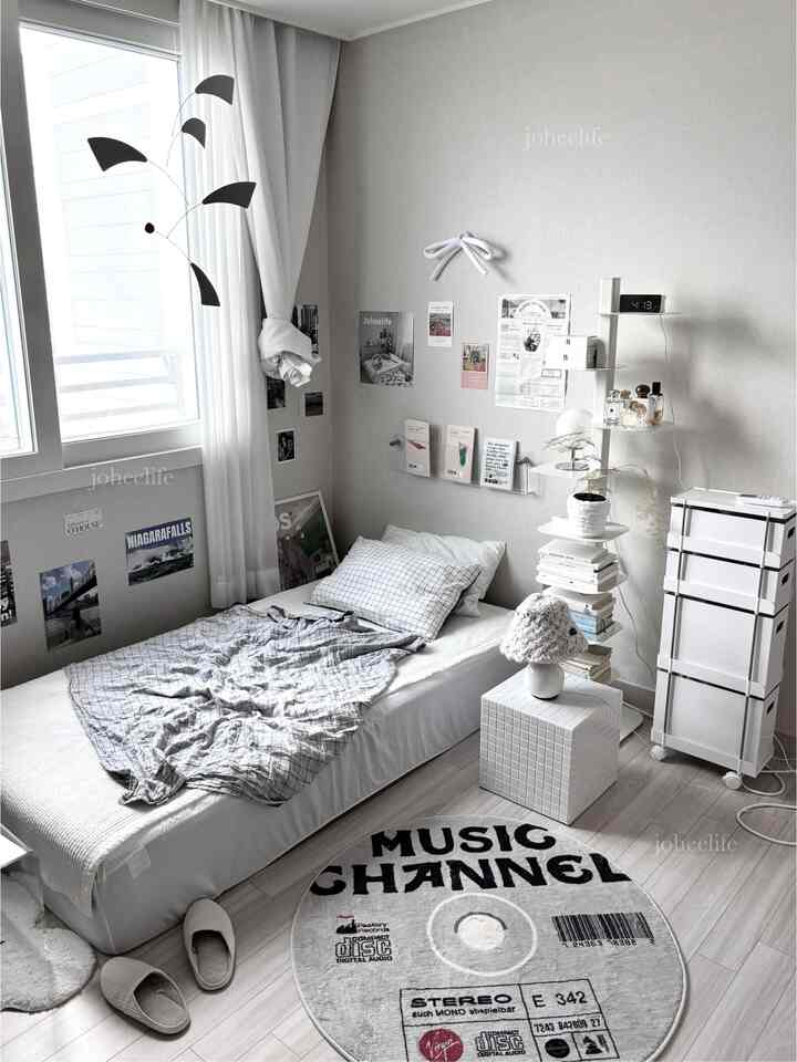 ホワイトとグレーのモノトーンの寝室、ロータイプのベッドと丸いラグ、壁には多彩な印刷物と収納棚が配置された整頓された空間