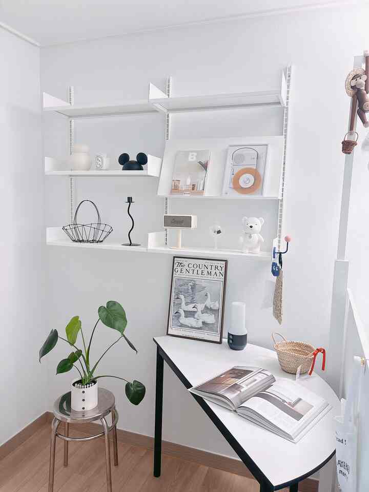 White-toned study space featuring white shelves, table, round stool, a plant, and decorative items in a simple modern style