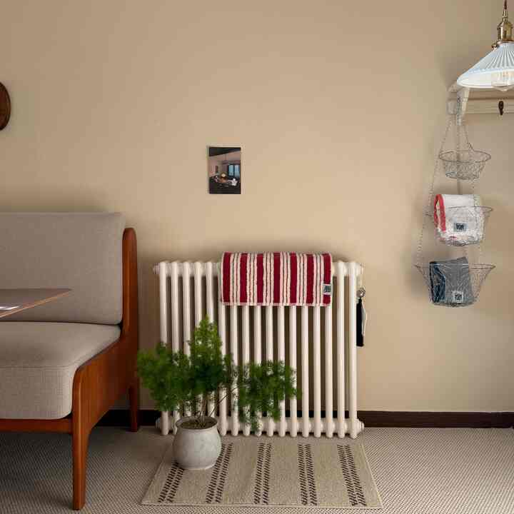 ベージュ色の壁を背景に、木製の肘掛けソファと白いラジエーターに赤いストライプのタオルがかかり、下にはラグと緑の観葉植物が配置されたナチュラルな印象のインテリア