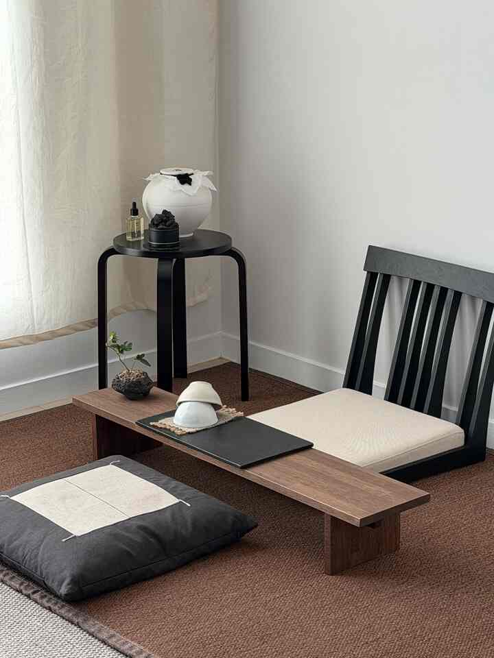 Beige-toned living space featuring a low wooden table, floor cushions, a black chair, and a small round side table, creating a natural minimalist interior