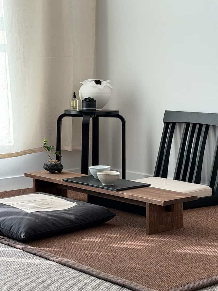 Natural-toned floor seating space featuring ivory curtains, black floor chair, walnut wood table, and Asian-style decor