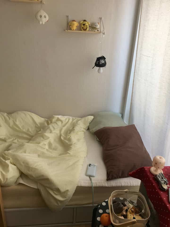 ベージュとイエローの落ち着いた色調の寝室、シングルベッドと枕、カーテン、小さな棚が配置された雰囲気のあるインテリア