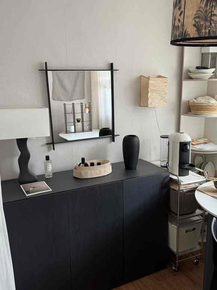 黒と白の色調のキッチン横の空間、黒い収納キャビネットとコーヒーマシン、ベージュの壁付け照明が配置された実用的でモダンな雰囲気のインテリア