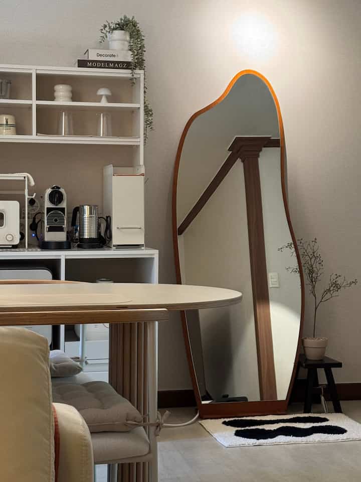 Modern kitchen space with white walls and wood-tone furniture, featuring a large irregular floor mirror and coffee machine in a simple interior