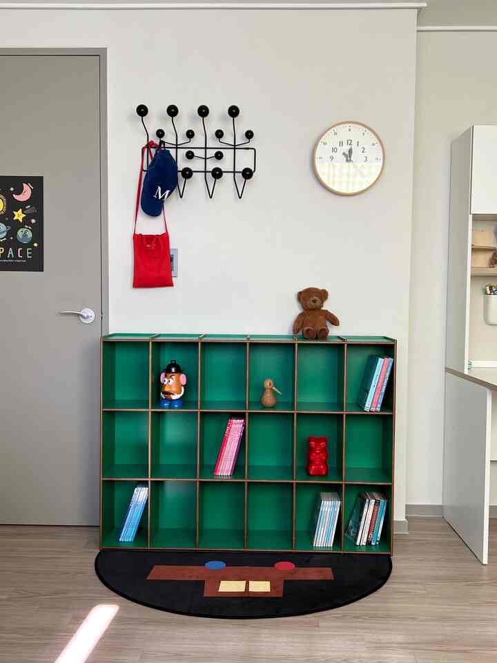 明るく整った子供部屋、中央に緑の本棚があり、壁には黒のハングイットオールと時計、床に可愛らしいラグが配置されている。