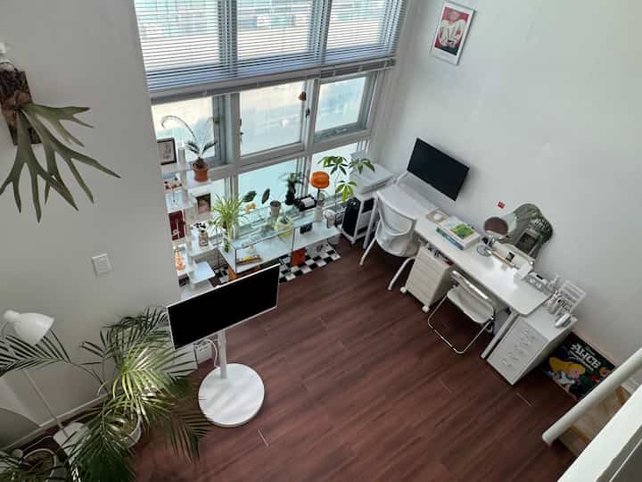 White walls and brown wood-tone floor define a loft-style home office featuring plants and a computer monitor, creating a clean workspace