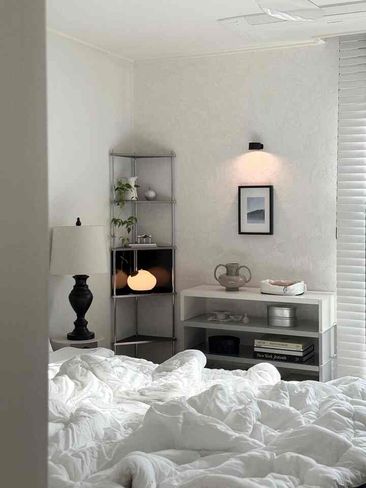 ホワイトトーンの寝室、中央に広げられた白い寝具と右側の金属製コーナー棚、壁付け照明が配置されたモダンな空間