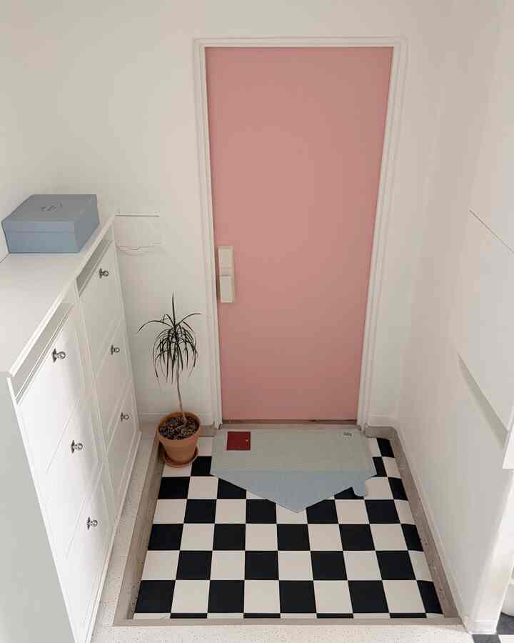 ピンクのドアと白い収納棚がある玄関、チェッカーフロアと青いマットが配置された雰囲気のある空間