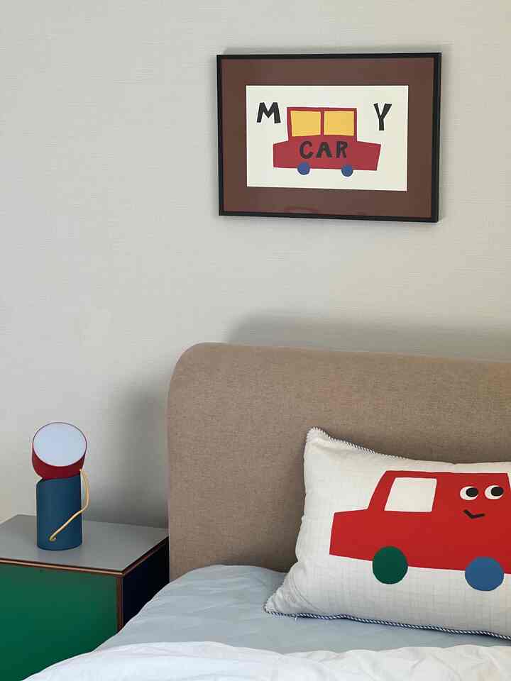 白い壁とベージュのベッドがある子供部屋、赤い車のイラスト入りクッションとポスターが雰囲気を演出