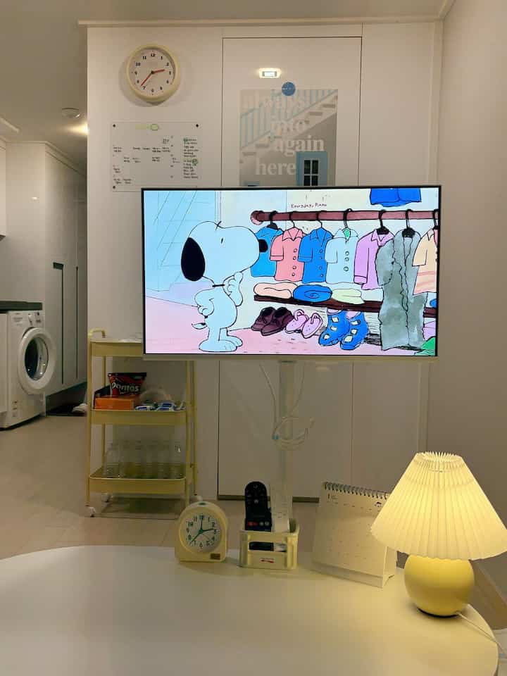 白を基調としたワンルームのリビング、壁時計とテーブルランプが配置された清潔で落ち着いた印象の空間