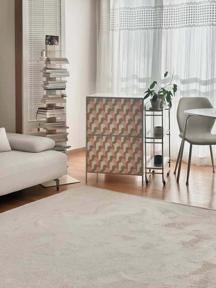 白とナチュラルな木目調のリビング、シンプルなソファと柄のキャビネット、透明なトロリーが配置された明るく落ち着いたインテリア