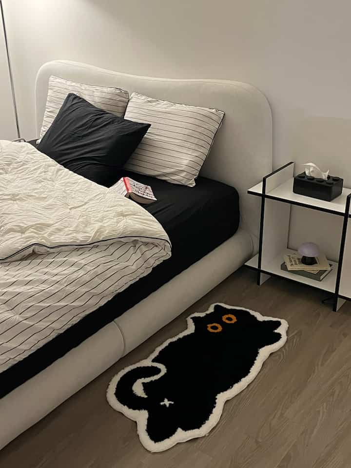 A cozy bedroom in white and black tones featuring a soft bed and a distinctive black cat-shaped rug