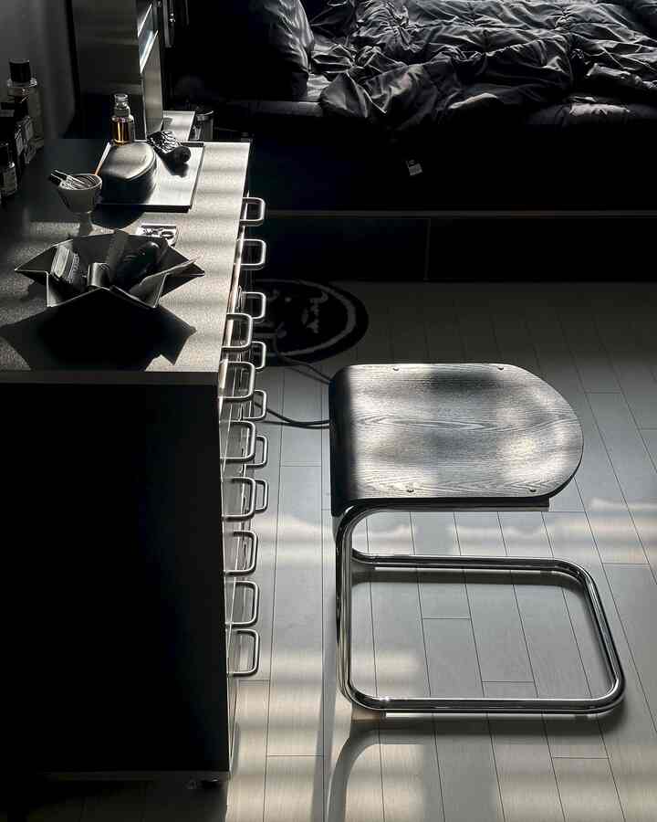 ブラックとグレーのトーンが特徴の寝室、中央にメタルフレームのスツール、左側にチェストが配置されたモダンな雰囲気のインテリア