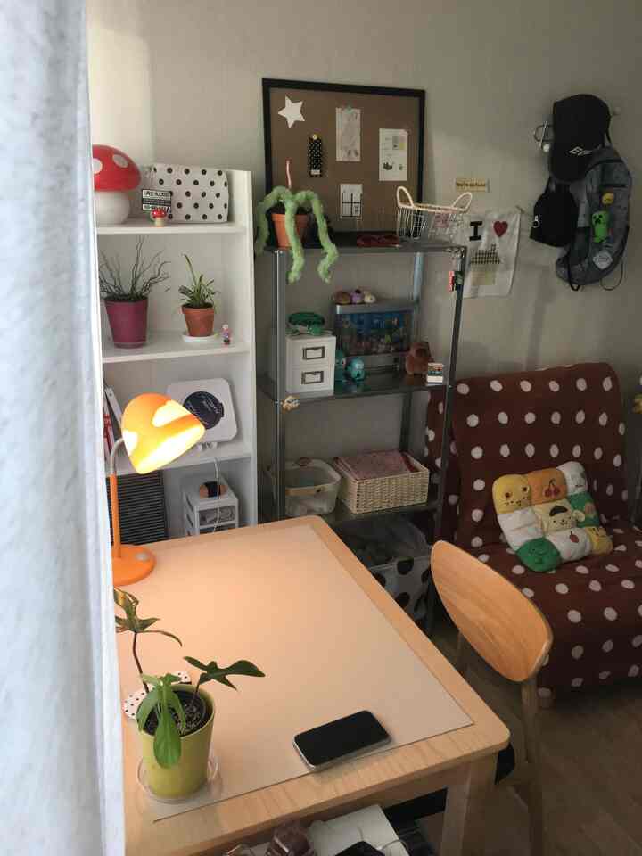 暖かいベージュ調の小さな部屋、左にテーブルクロスの敷かれたデスク、右にドット柄のソファベッド、棚や植物が配置された雰囲気のあるホームオフィス空間